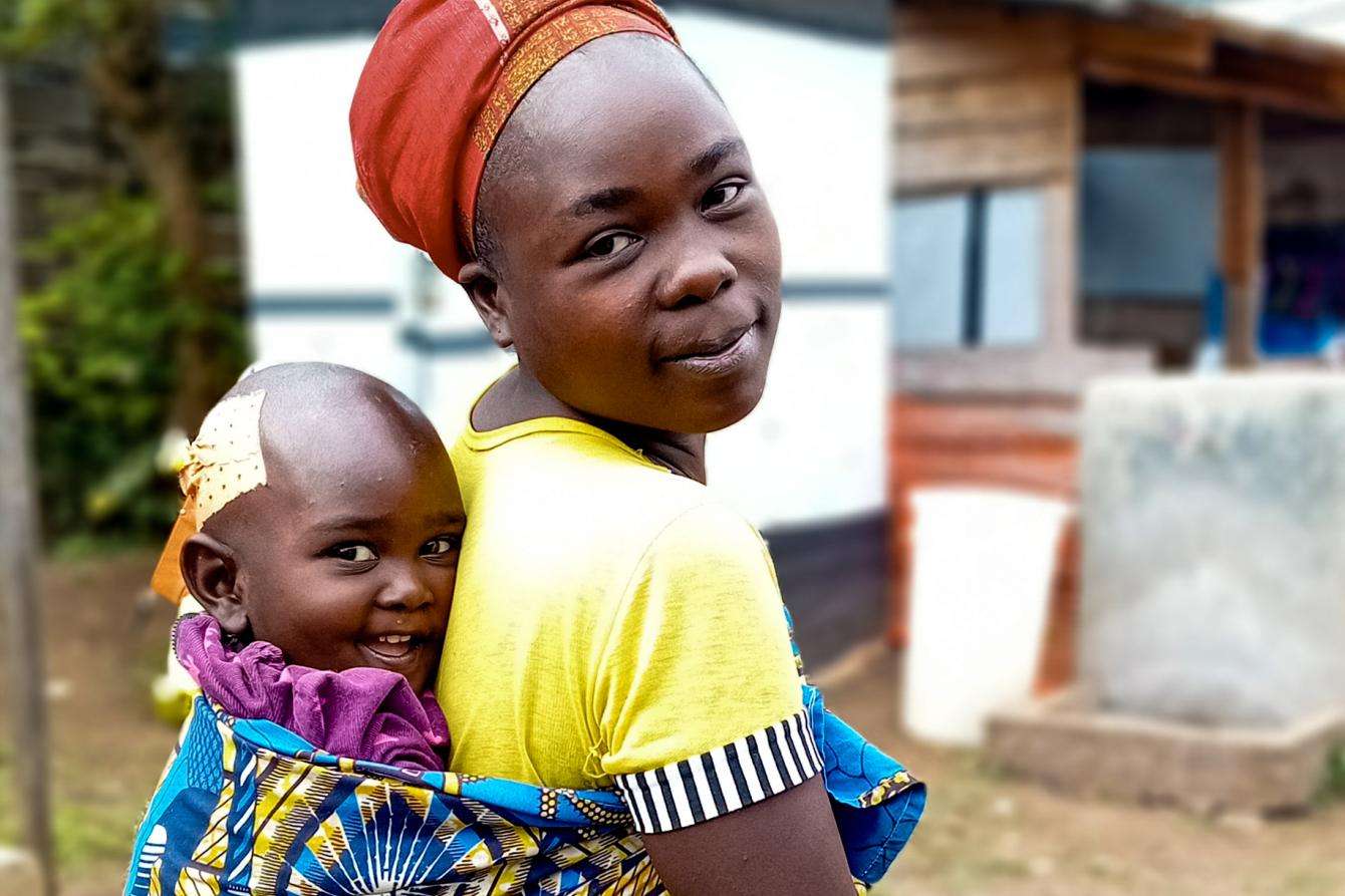 A woman in colorful clothing (a teal dress and red headdress) is standing outside, smiling holding her child. 